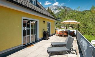 A sunny terrace with loungers and a dining table under a sun umbrella. In the background, green trees and mountains are visible.