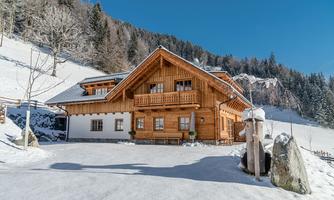 A beautiful wooden house in a snowy landscape. The surroundings are filled with tall trees and a clear blue sky.