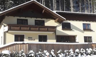 A snow-covered house in the mountains with a beautiful balcony. The surroundings are surrounded by tall, green trees.