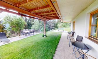 An inviting outdoor area with grass, a covered terrace, and comfortable seating. In the background, trees and a tranquil landscape are visible.