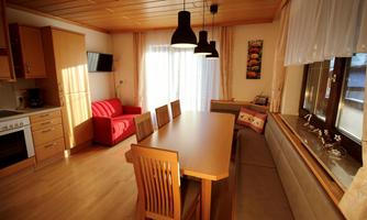 A bright kitchen with a large dining table and wooden chairs. There is a cozy red sofa and plenty of daylight through the windows.
