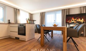 A modern living room with an open kitchen and a cozy dining area. The room is bright and inviting, with a fireplace in the background.