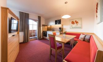 A cozy apartment with a dining area and a kitchenette. The room features inviting red furniture and plenty of natural light.