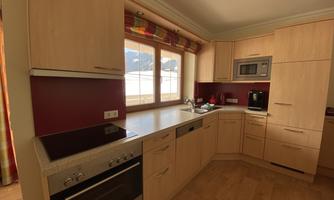 A modern kitchen with wooden cabinets and a red accent on the wall. The room is bright and inviting with large windows.