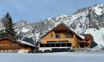 A wooden house in the mountains with snow-covered areas. Imposing rocks are visible in the background.