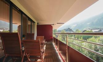 A modern balcony with wooden chairs and a table. In the background, green hills and trees are visible.