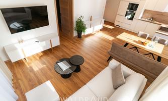 A modern living room with a bright wooden floor and a white sofa. In the background, an open kitchen with a dining table and chairs is visible.