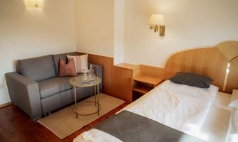 A cozy bedroom with a gray sofa and a small side table. The room has light walls and a double bed with plain bedding.