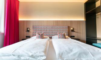 A modern hotel room with two single beds and colorful cushions. Red curtains and stylish walls create an inviting atmosphere.