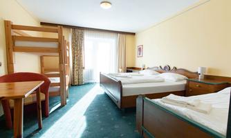 A simple hotel room with two beds and a bunk bed. A table and chairs are available in the room, with plenty of natural light from the window side.