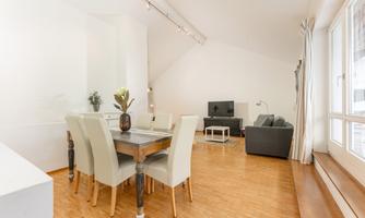 A modern living room with a dining area and a comfortable couch. The room is bright and inviting, with wooden floors and large windows.