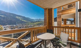 A balcony with a table and chairs that offers a magnificent view of the mountains. The sun is shining and the sky is clear.