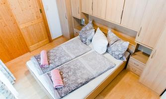 A cozy bedroom with a double bed and two pillows. The walls are made of light wood, and a wooden wardrobe can be seen in the background.