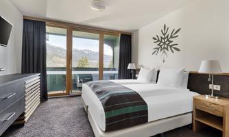 A modern hotel room with a double bed and a balcony. The large windows offer a view of the mountains.
