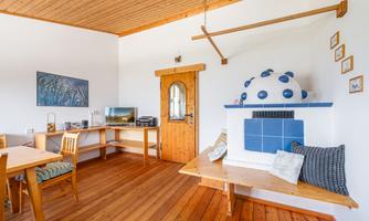 A cozy living space with wooden floors and clean lines. There is a small table, chairs, and a charming tiled stove.