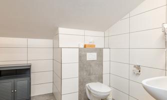 A modern bathroom with white tiles and a simple design. There is a toilet, a sink, and a small chest of drawers.