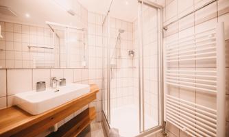 A modern bathroom with a shower and a wooden sink. The walls are tiled, and there is plenty of daylight.