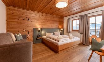 A cozy hotel room with wood paneling and a large window. The room features a double bed, a seating area, and plenty of daylight.