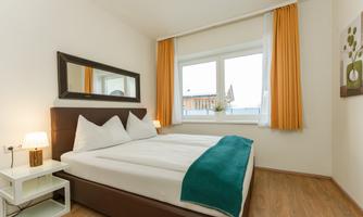 A cozy bedroom with a double bed and white pillows. Large windows with curtains let in plenty of daylight.