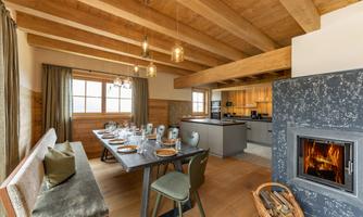An elegant dining area with a large table, surrounded by comfortable chairs. In the background, a modern kitchen and a fireplace are visible, creating a cozy atmosphere.