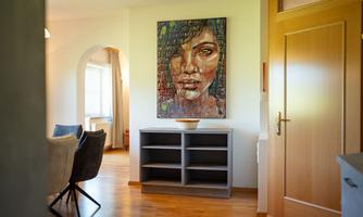 A bright living space with a wooden floor and a large painting of a woman on the wall. In the foreground are a couple of chairs and a shelf.