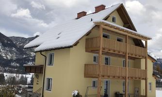 A yellow house with a snow-covered roof and balcony. In the background, mountains and a cloudy sky can be seen.