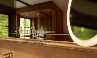A modern bathroom with an elegant sink and a large mirror. In the background, wooden details and windows with a view of greenery can be seen.