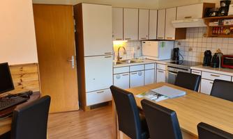 A modern kitchen with a large table and chairs. In the background, various kitchen appliances and a refrigerator can be seen.