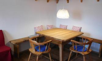 A cozy dining area with a wood-colored table and chairs. The wall is decorated with shelves and a lamp provides warm light.