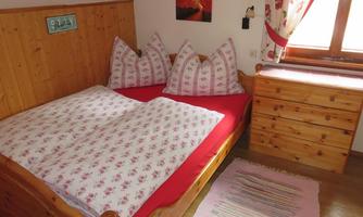 A cozy bedroom with a wooden bed and red bedding. On the wall, there's a small picture, and a wooden wardrobe stands next to the window.