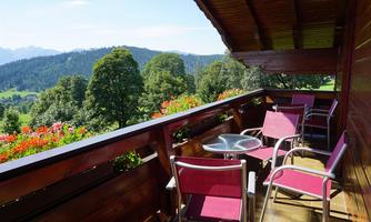 A balcony with modern seating and a table. In the background, beautiful green mountains and trees can be seen.