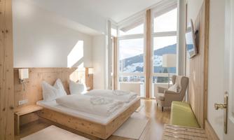 A bright room with a double bed and wooden paneling. Large windows offer a view of the mountains.