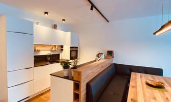 A modern kitchen with white cabinets and a dark countertop. The dining area consists of a wooden bench and a table.