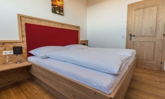 A cozy bedroom with a double bed and wooden furniture. The walls are painted light and there is a window with natural light.