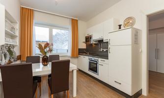 A modern kitchen with a dining area. The room has large windows and wooden flooring.