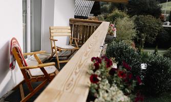 A cozy balcony with a wooden chair and colorful flower boxes. In the background, a green landscape is visible.