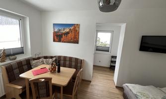 A cozy living area with a dining table, chairs, and a sectional sofa. A picture of an autumn landscape hangs on the wall.