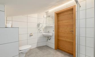 A modern bathroom with white tiles and a wooden steel door. There is a shower, a sink, and a toilet.