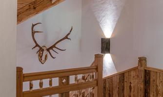 A rustic staircase with wooden railing and ceiling beams. A deer antler hangs on the wall, emphasizing the alpine style.