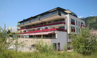A modern apartment building with balconies and red accents. It is surrounded by green vegetation and a blue sky.