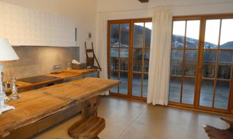 A modern kitchen with a rustic wooden table and bar stools. Large windows offer a view of the surrounding landscape.