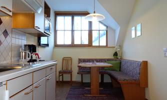 A cozy kitchen with wooden cabinets and windows that let in daylight. There is a dining area with a table and a bench.