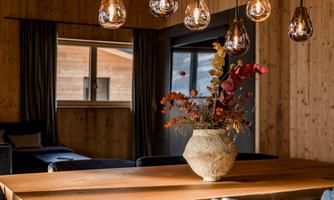 A modern designed dining area with a large wooden table and elegant chairs. Stylish lamps hang above the table, and a decorative vase with flowers stands in the center.