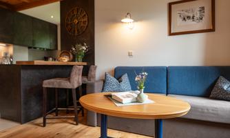 A cozy living room with a blue sofa and a round wooden table. In the background, a modern kitchen and a wall decoration can be seen.