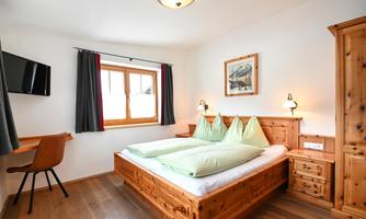 A cozy bedroom with wooden furniture and a double bed. The room has a window with curtains and a television on the wall.
