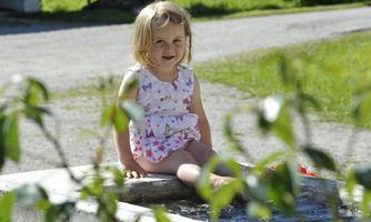 A small girl is sitting at the edge of a water feature and smiling. In the background, you can see a green meadow and trees.
