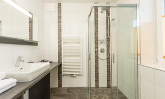 A modern bathroom with a glass shower and a large sink. The walls are light tiled and there is a towel radiator.