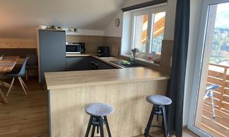 A modern kitchen with a wooden bar and two stools. Large windows provide a beautiful view outside.