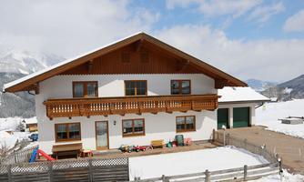 A traditional two-story house in the snow, surrounded by mountains. The facade is white with wooden decorations and a terrace.