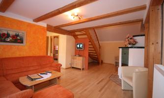 A cozy living room with an orange sofa and wooden beams. In the corner, there is a staircase and a television.